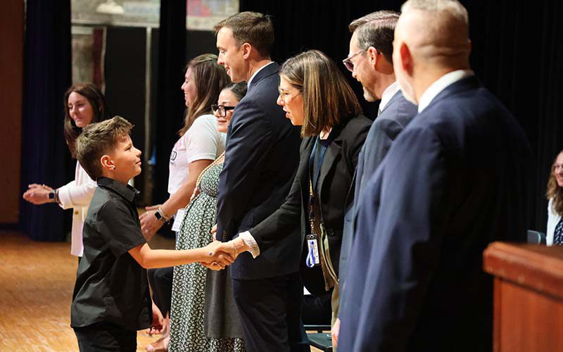 A fifth-grade boy shakes hands with a woman on a stage.