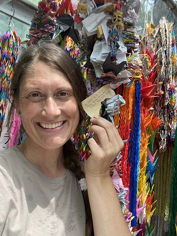 A woman smiles as she holds up a note that says Pine Bush Elementary School, NY, USA. Behind her are thousands of paper cranes.