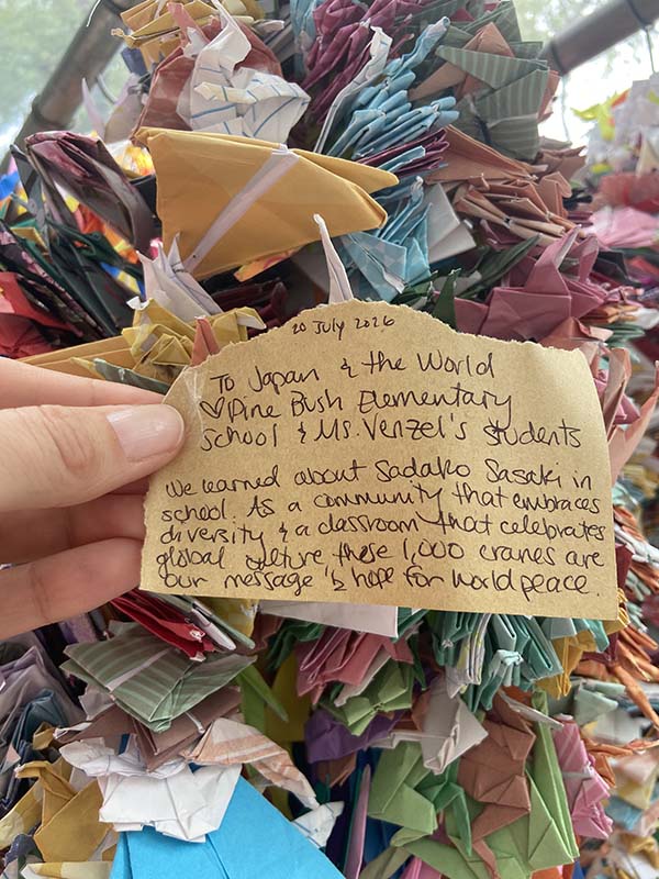 In the background are paper cranes. The note in the forefront says We learned about Sadako Sasaki in school. As a community that embraces diversity and a classroom that celebrates global culture, these 1,000 cranes are our message of hope for world peace."