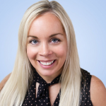 A woman with long blonde hair, wearing a black shirt with gold dots, smiles.