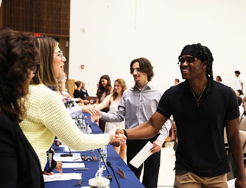 A high school senior shakes hands with a woman . Behind him are other students shaking hands with others adults.