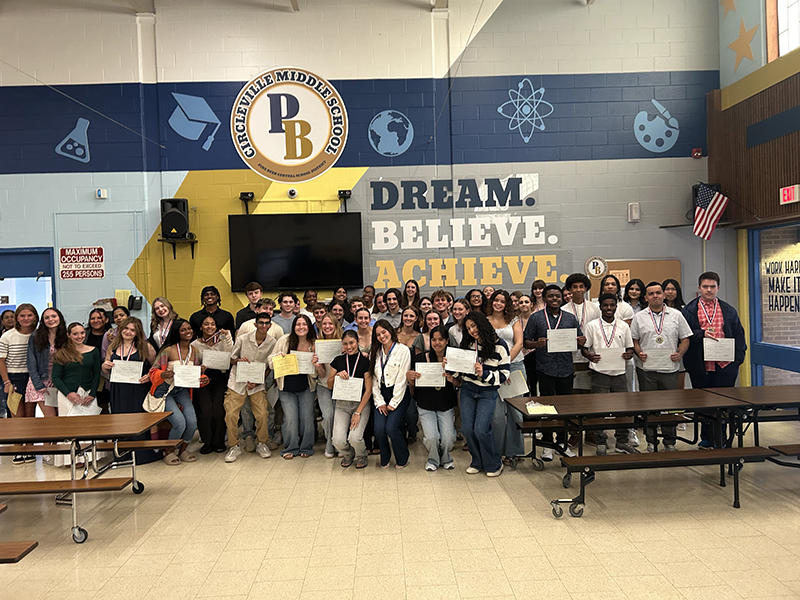 A large group of high school students stand together and smile, about 70 of them. They are in front of a wall painted in blue, gold and white that says Dream. Believe. Achieve.