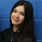 A young woman with long dark hair smiles. She is in front of a blue background.