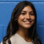 A young woman with long dark hair smiles. She is wearing a tan shirt and is in front of a blue background.