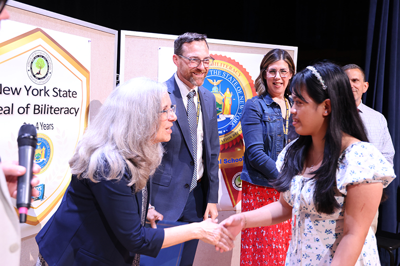 A woman shakes hands with a young woman on a stage. Others looks on and smile.