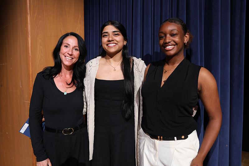A woman stands on the left and two high school seniors are next to her. All are smiling.