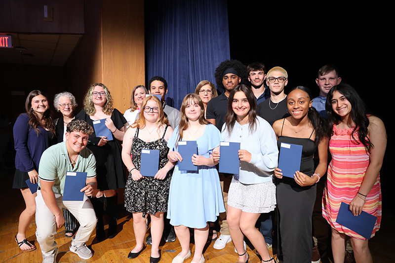 A group of 16 high school seniors stand together holding certificates on a stage.