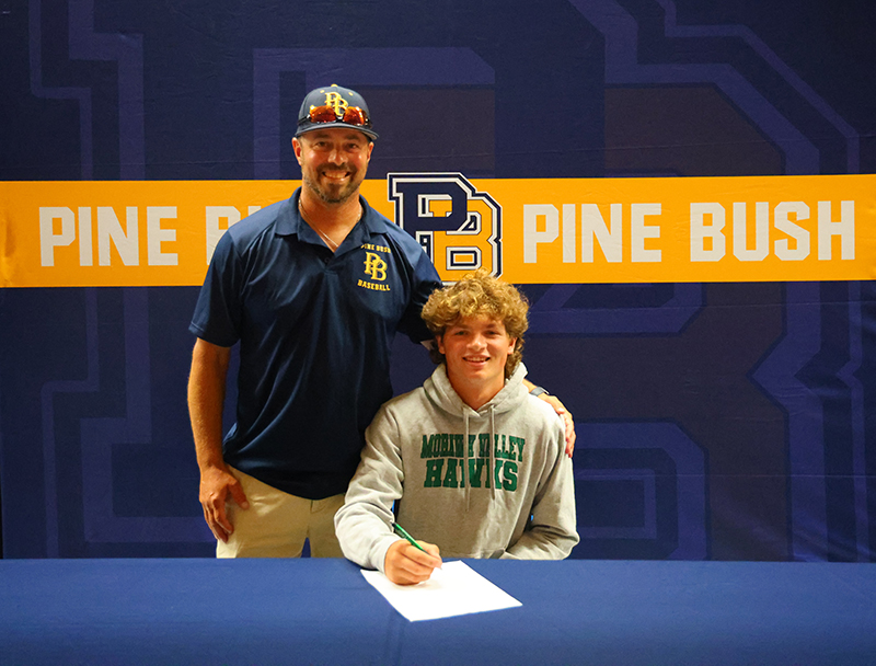 A man stands next to a young man who is sitting. They are both smiling and there is a banner behind them that says Pine Bush.