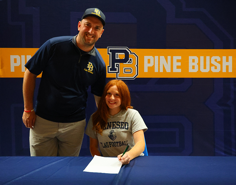 A high school girl wearing a Geneseo flag football shirt sits at a table an smiles. There is a man next to her smiling, wearing a Pine Bush Flag Football shirt and hat. Behind them is a backdrop in blue and gold that says Pine Bush.