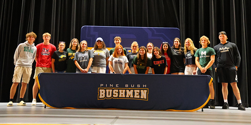 a group of 17 high school kids, all wearin gdifferent college tshirts, stands together behind a table that says Pine Bush Bushmen.