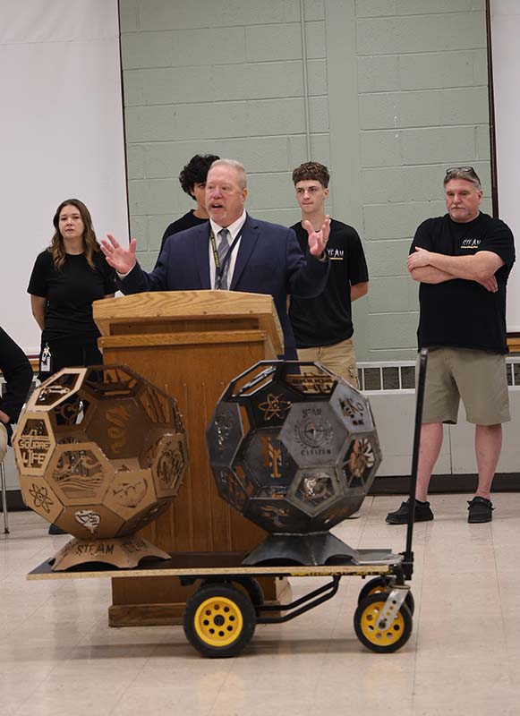 A man stands at a podium speaking with projects in front of him.