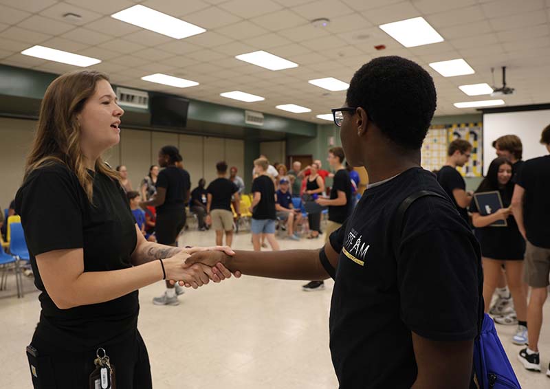 A young man shakes hands with a woman. There are many people in the background.