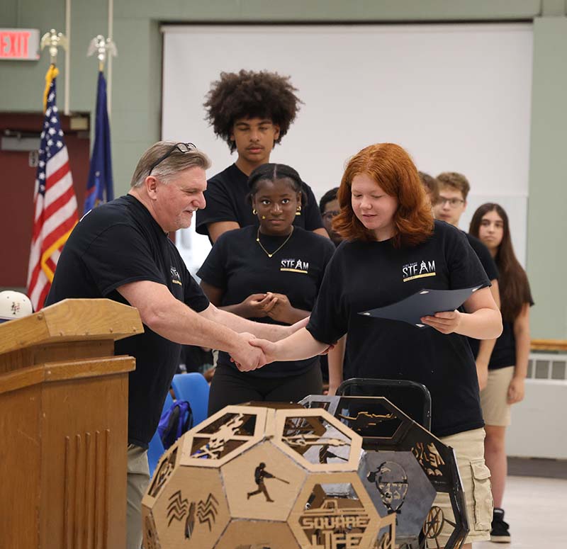 A high school girl shakes hands with a man by a podium. There are other high school kids behind her.