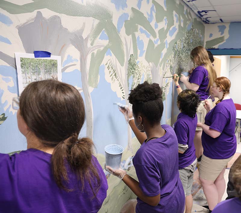 A group of five high school students paint a mural on a large wall. The painting is of a forest.