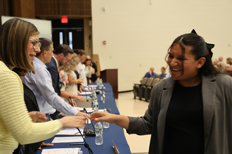 A high school student shakes hands with a woman behind a table.