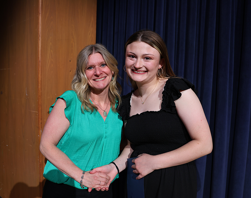 A woman on the left and a high school senior girl stand together and smile.