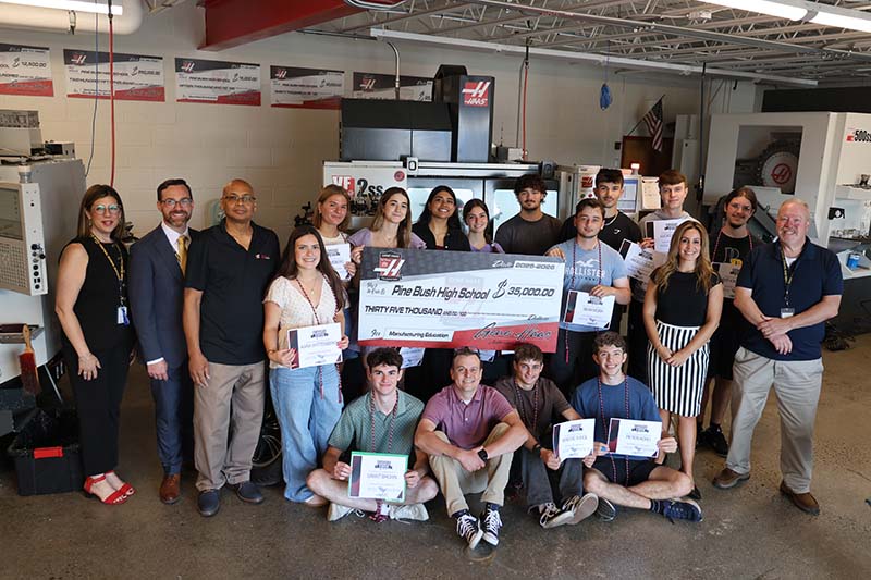 A group of 13 high school students and five adults stand together. Several of the students are holding an oversized check.