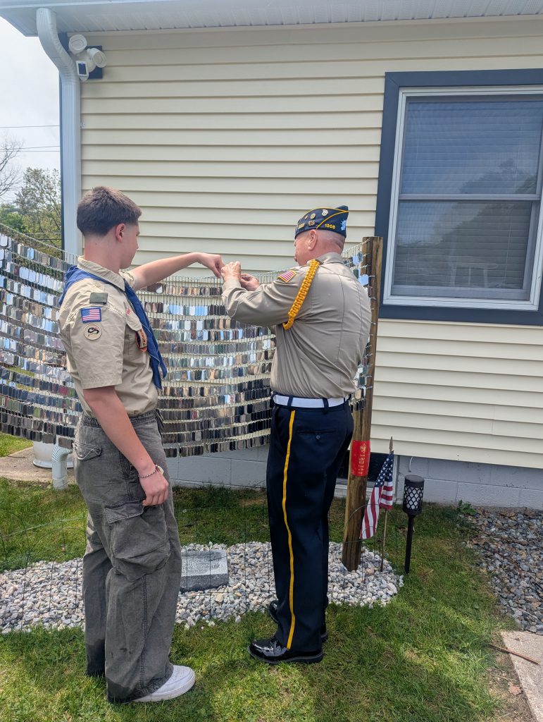 A man and a young man, both in uniform, hang dog tags on on a memorial