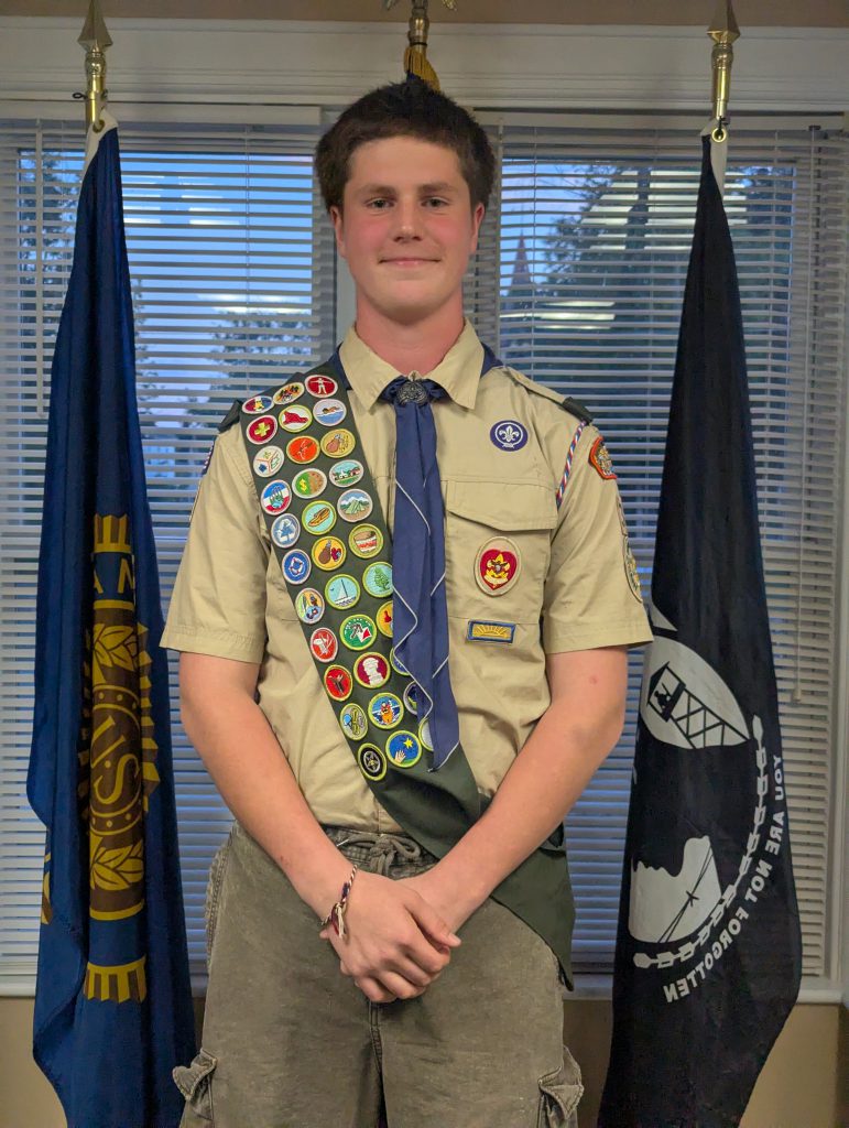 A young man smiles. He is standing with a boy scout uniform on with many merit badges.