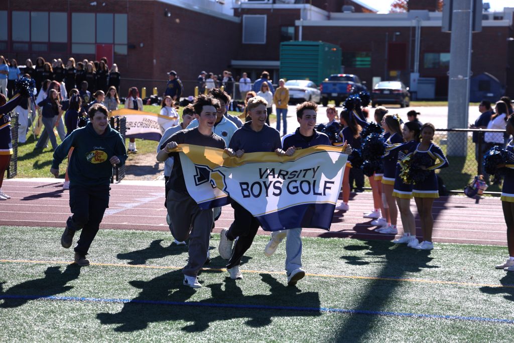 high schoolers running out on football field holding a banner that says varsity boys golf